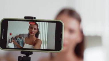 Genç sosyal medyum kadın moda videosu yapıyor. Blogcu kameraya selfie çubuğu ve ışık halkası kullanarak gülümserken, aynı zamanda ikna edici çevrimiçi kıyafetlerin izleyicilere veya takipçilere vlog satmasını sağlıyor. Düzenle