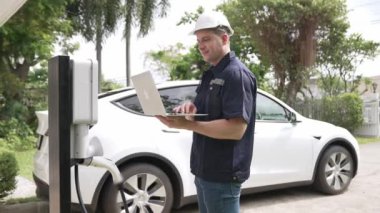 Evde EV şarj istasyonu kurulumu üzerinde çalışan nitelikli teknisyen, EV için dizüstü bilgisayarla şarj sisteminde sorun çıkarma ve yapılandırma ayarları yapıyor. Titiz