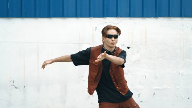 Beyaz arka planda break dans pratiği yapan yakışıklı bir sokak dansçısı. Hip hop tarzı giyinen sporcu, mavi arka planlı bir sokakta müzik eşliğinde ayak sesleri çıkarıyor. Açık hava sporu 2024 hiphop..