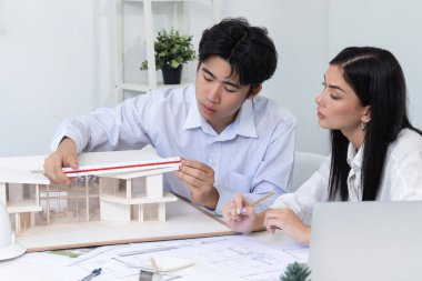 Profesyonel Asyalı erkek mimar ev modelinin uzunluğunu ölçmek için cetvel kullanırken, beyaz tenli genç meslektaşı ev modeliyle toplantı masasındaki verileri analiz etmek için dizüstü bilgisayar kullanıyor. Tertemiz..