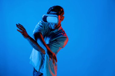 Yakışıklı, dans eden ve müziğe hareket eden bir adam. VR gözlüğü ve neon ışıklı arka plan kullanarak. Beyaz erkek oyuncu, teknolojik yenilikler giyerek pop müzik dinlerken ayakta duruyor. Sapma.