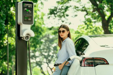 Yolculuk sırasında doğal orman ya da milli parktaki elektrikli araba için şarj olan genç bir kadın. Tatil ve tatil boyunca çevre dostu seyahat. Yükselt