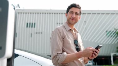 Ağır çekimde çalışan adam, alışveriş merkezinin otoparkındaki kamu EV şarj istasyonunda elektrik için akıllı telefon kullanıyor. EV aracıyla modern çevre ve sürdürülebilir kentsel yaşam tarzı