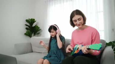 Asyalı tatlı kız kulaklıkla müzik dinlerken güzel anne de akustik müzik öğretirken ukulele çalıyor. Sevimli çocuk enstrüman öğreniyor. Pedagoji.