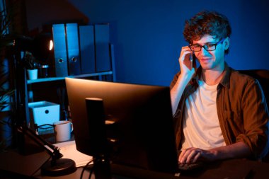 Genç iş adamı akıllı telefondan müşteriyi ya da iş arkadaşını arıyor, neon modern ofisteki yaratıcı sunum surround çalışma masasının bilgisayar ekranına bakıyor. Yeni proje kavramı. Toplayıcı.