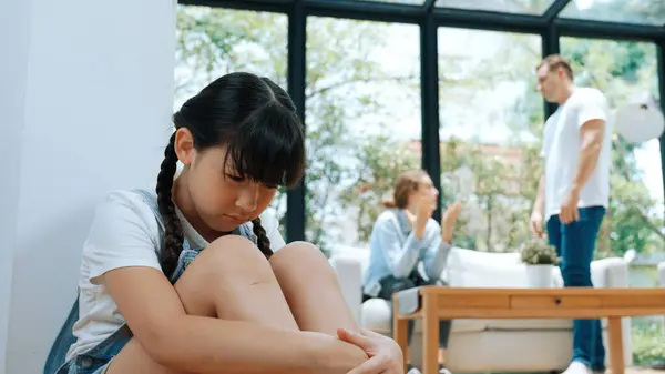 stock image Stressed and unhappy young girl huddle in corner, cover her ears blocking sound of her parent arguing in background. Domestic violence at home and traumatic childhood develop to depression. Synchronos