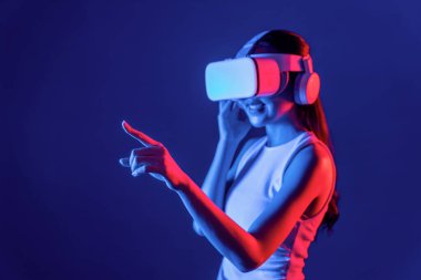 Siberpunk neon ışıklı cihazlarla çevrili zeki bir kadın metaevrene bağlı VR kulaklık takıyor, gelecekçi siber uzay teknolojisi. Sanal objeyi işaret eden bir kadın. Halüsinasyon.