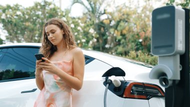 Genç bir kadın doğadaki EV şarj istasyonunda elektrik için akıllı telefon kullanıyor. Modern çevre ve EV araçlı sürdürülebilir otomobil taşıma yaşam tarzı. Eşzamanlama