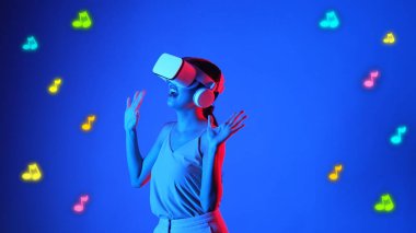 Müzik ve teknoloji kavramının mavi arka planında, stüdyo dinamik ışıklandırmasında VR kulaklıkla müzik notaları ve modern tasarım kullanarak eğlence şarkıları dinleyen ve dans eden gülümseyen bir kadın. Halüsinasyon.