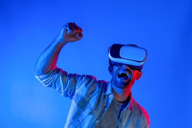 Sanal dünyada spor müsabakalarını izleyen mutlu adam neon ışıklı arka planda VR kulaklık takıyor. Yakışıklı genç, gelecekteki yenilik teknolojisine gözlük kullanarak giriyor. Sapma.