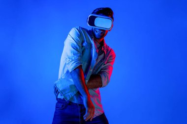Yakışıklı, dans eden ve müziğe hareket eden bir adam. VR gözlüğü ve neon ışıklı arka plan kullanarak. Beyaz erkek oyuncu, teknolojik yenilikler giyerek pop müzik dinlerken ayakta duruyor. Sapma.
