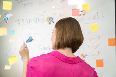 Güzel iş kadını, zihin haritası ve yapışkan notla yaratıcı pazarlama stratejisini düşünürken cam panoya etiket yapıştırıyor. Yaratıcı iş toplantısı konsepti. Arkadan bak. Tertemiz..