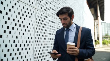 Akıllı yönetici cep telefonuna bakıyor ve takım elbise giyerken sokakta yürüyor. Yetenekli yatırımcı cep telefonuyla satışları kontrol ediyor ya da elinde kahve fincanı tutarken çalışıyor. Beyaz arka plan. Sevinçli..