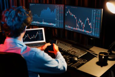 Akıllı telefonu elinde tutan genç yatırımcı hisse senedi tüccarı bilgisayardaki dinamik döviz kuru verileriyle karşılaştırıyor ve güncel pazar grafiğinde Neon Light Modern Ofis 'te gerçek zamanlı olarak. Toplayıcı.