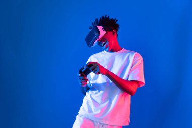 VR kullanan Amerikalı Afrikalı oyun oyuncusu joystick 'i kontrol eden hologram izole edilmiş mavi neon ışıklı duvar ekranı dijital fütüristik teknoloji sanal gerçeklik dünyasına bağlanıyor. Entrika.