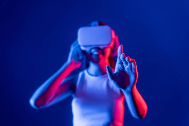 Çevresi siberpunk neon ışıklarıyla çevrili zeki bir kadın metaevrene bağlı VR kulaklık takıyor, gelecekçi siber uzay topluluğu teknolojisi. Sanal objeye el ile dokunan kadın. Halüsinasyon.