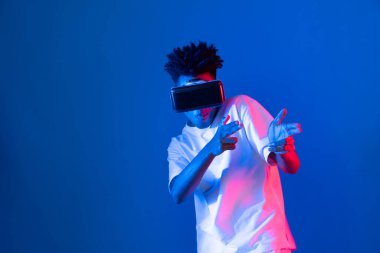 VR kullanan genç Afrikalı Amerikalı oyuncu sanal gerçeklik dünyasını dijital fütüristik teknolojiye bağlayan mavi neon ışıklandırma duvarında 3 boyutlu sanal silah avcısını kullanıyor. Entrika.