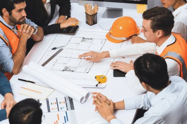 Mimari proje üzerinde birlikte çalışan çeşitli inşaat mühendisi ve müvekkil grupları, inşaat planını gözden geçirip toplantı masasında plan oluşturuyorlar. Tedbirli