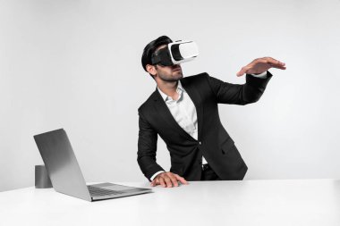 Profesyonel proje yöneticisi dizüstü bilgisayar başında otururken VR gözlüğü kullanıyor. Görsel gerçeklik kulaklığı takan yetenekli bir iş adamı metaevren ya da görsel dünyayı birbirine bağlıyor. Yenilik. Sapma.