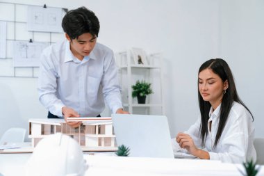 Profesyonel Asyalı erkek mimar ev modelinin uzunluğunu ölçmek için cetvel kullanırken, beyaz tenli genç meslektaşı ev modeliyle toplantı masasındaki verileri analiz etmek için dizüstü bilgisayar kullanıyor. Tertemiz..