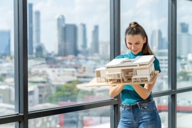 Güzel genç mimar mühendisi ev modelini incelerken mimari modeli elinde tutuyor. Şehir manzaralı pencerenin yanında dururken profesyonel iç mimar ev inşaatını kontrol ediyor. İzleme.