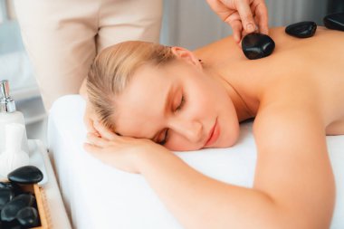 Lüks tatil köyündeki kaplıcalarda sıcak taş masajı. Gün ışığı sükunet ortamında, mutlu kadın müşteri bazalt taş masajından zevk alıyor. Rahatlatıcı bir sıcaklıkla vücudun üzerinde süzülüyor. Sessiz.