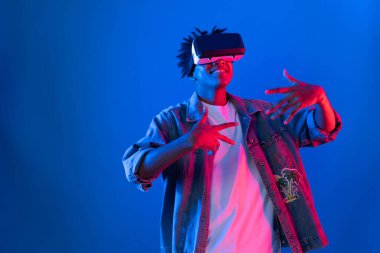 Gülümseyen Afrikalı genç Amerikalı sanal gerçekliği dijital fütüristik teknolojiye bağlayan pembe mavi neon duvarda hologram oyunlarıyla VR dansına bakıyor. Entrika.