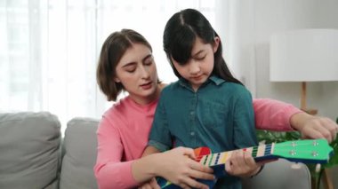Kafkasyalı bir anne evde akustik müzik öğretirken ukulele çalan mutlu kız. Sevimli çocuk enstrüman öğreniyor. Kafkasyalı mutlu anne ve kız birlikte vakit geçiriyorlar. Pedagoji.