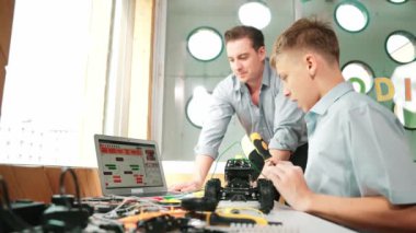 Kafkas öğretmeni dizüstü bilgisayarı işaret ederek ve programlama sistemi veya kodlama için tavsiye verirken zeki liseli çocuk STEM sınıfında robotu tamir etmek için ekipmanları dinliyor ve kullanıyor.