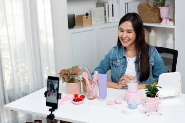 Kozmetik ürünlerini ve cilt bakım ürünlerini inceleyen genç ve güzel bir Asyalı kız sosyal medyada canlı yayında satışa sunulan yeni koleksiyon promosyonları ile güzellik ürünlerini çevreleyen bulanık telefon ekranı. Taktik.