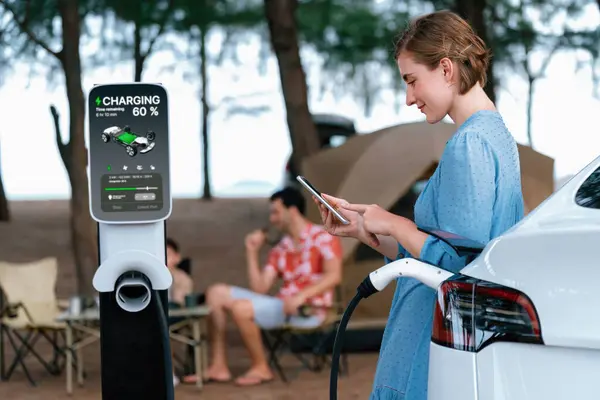 stock image Outdoor adventure and family vacation camping at sea travel by eco friendly car. Woman or mother check cars battery with smartphone while charging EV car frin charging station in campsite. Perpetual