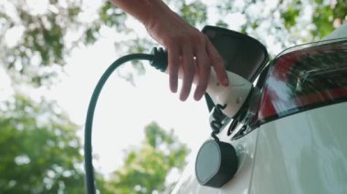 Yavaş çekim adamı, yaz mevsiminde açık yeşil yay yeşilliğindeki şarj istasyonundan elektrikli araba aküsünü şarj etmek için EV şarj aleti taktı. Yeniden şarj edilebilir elektrikli araç için sürdürülebilir enerji