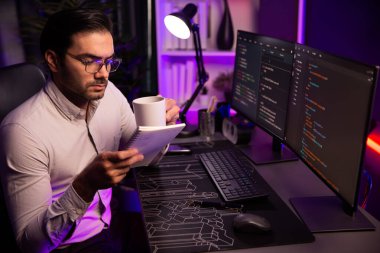 Akıllı bilişim geliştiricisi, bilgisayar ekranındaki yazılım geliştirme kodlaması üzerine kahveyle birlikte çalışıyor, en yeni uygulama projesi programı ile birlikte neon ışıklı siber ofiste çevrimiçi web sitesi verilerini güncelliyor. Tahmin et..