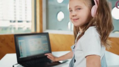 Masaya dizüstü bilgisayarı yerleştirip kameraya el sallayan gülümseyen kız. BTMM sınıfında dizüstü bilgisayar programlama ya da kodlama programı yaparken kulaklık takan bir çocuk gülümsüyor. Etkinlik.