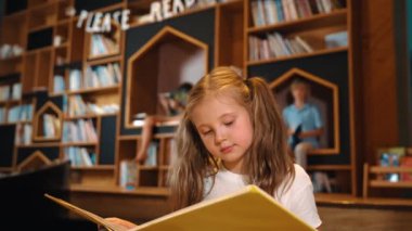 Genç, zeki, beyaz bir kız kütüphanede otururken kitap okuyor. Zeki çocuk öğrenme, okuma, kütüphanede kitap açma. Etkileyici çocuk geçmişi bulanık sayfayı çeviriyor. Etkinlik.