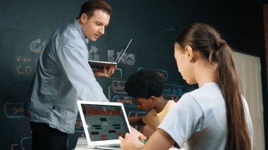 Afrikalı erkek manken tamir ederken beyaz kız programlama ya da kodlama sistemi. Öğretmen elinde dizüstü bilgisayar tutarken çeşitli çocukların web geliştirme programı yazmalarına bakıyor. Yenilik eğitimi. Öğretim.