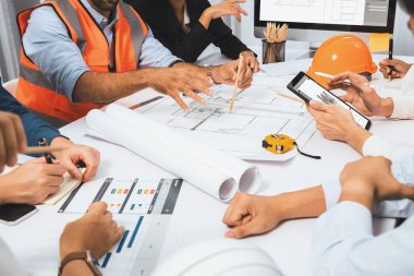 Mimari proje üzerinde birlikte çalışan çeşitli inşaat mühendisi ve müvekkil grupları, inşaat planını gözden geçirip toplantı masasında plan oluşturuyorlar. Tedbirli