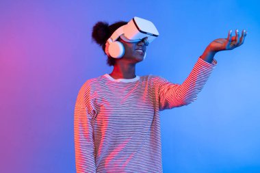 VR kullanan gülümseyen Afrikalı kadın meta-evren çiçekleri aracılığıyla güzel kokuyor ve kelebek böceği hologramı canlı pembe ışıklı arka plan teknolojisi 3D siber uzay. Entrika.