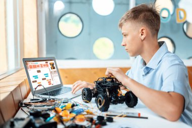 Liseli çocuk ekranda kod izliyor ve STEM sınıfında teknoloji öğrenirken robotik araba tutuyor. Masaya dizüstü bilgisayar, kumanda, elektrik kablosu, şarj aleti ve robot araç koyun. Öğretim.