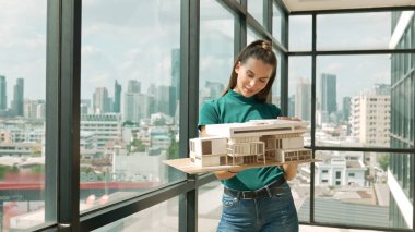Genç akıllı mimar mühendisi ev modelini incelerken mimari modeli elinde tutuyor. Şehir manzaralı pencerenin yanında dururken profesyonel iç mimar ev inşaatını kontrol ediyor. İzleme