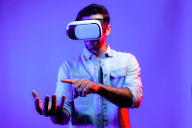 Sanal gerçeklik gözlüklü, sanal gerçeklikte hologram görüntüsü olan beyaz, zeki bir adam. Neon ışıklı arka planda gözlük ve kulaklık kullanarak bakıp işaret eden mutlu bir insan. Teknoloji. Sapma.