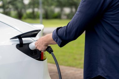 Açık yeşil şehir parkı manzaralı EV şarj istasyonundan EV elektrikli araçları şarj eden adam. Çevre dostu şehir ulaşımı ve çevre dostu EV araba seyahati. Yükselt