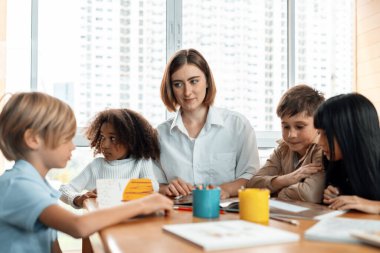 Öğretmen sınıftaki kitaplardan ders çıkarmayı öğretiyor, çocuklar mutlu. Bazıları öğretmenine soruyor, mavi gömlekli bir çocuk tek başına kitap okuyor ve bir kız resim çiziyor. Etkinlik.
