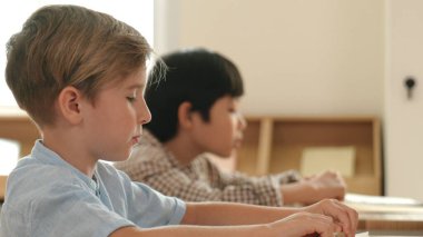 Beyaz tenli ilkokul öğrencisinin tahtada otururken bir nota bakması ve soruya cevap vermesi. Çocuklar derse katılırken genç öğretmen sınıf çalışmasını ya da sınavı açıklar. Pedagoji.