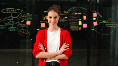 Profesyonel genç yönetici, silah zoruyla kameraya bakıyor. Cam duvarın önünde zihin haritası ve yapışkan notlarla dikilen yetenekli bir işçi. İzleme