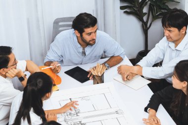 Mimari proje üzerinde birlikte çalışan çeşitli inşaat mühendisi ve müvekkil grupları, inşaat planını gözden geçirip toplantı masasında plan oluşturuyorlar. Tedbirli