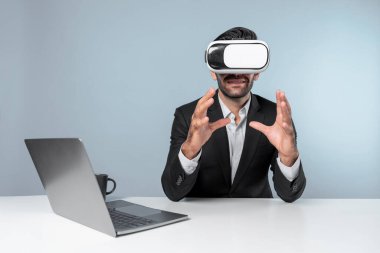 Profesyonel proje yöneticisi dizüstü bilgisayar başında otururken VR gözlüğü kullanıyor. Görsel gerçeklik kulaklığı takan yetenekli bir iş adamı metaevren ya da görsel dünyayı birbirine bağlıyor. Yenilik. Sapma.