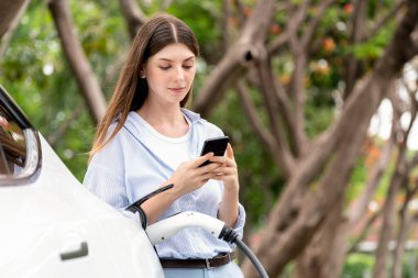 Genç bir kadın ulusal park veya sonbahar tatili gezisi sırasında EV şarj istasyonundan elektrik aküsü almak için internet üzerinden akıllı telefon bankacılık uygulaması kullanıyor. Yükselt
