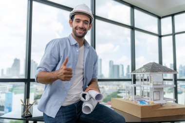 Modern ev projesini düşünen profesyonel mimar mühendisinin portresi güvenlik kaskı ve günlük kıyafetler giyerken. Akıllı iç mimar ev modelinin yanında proje planları tutuyor. İzleme.