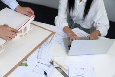 Profesyonel Asyalı erkek mimar ev modelinin uzunluğunu ölçmek için cetvel kullanırken, beyaz tenli genç meslektaşı ev modeliyle toplantı masasındaki verileri analiz etmek için dizüstü bilgisayar kullanıyor. Tertemiz..